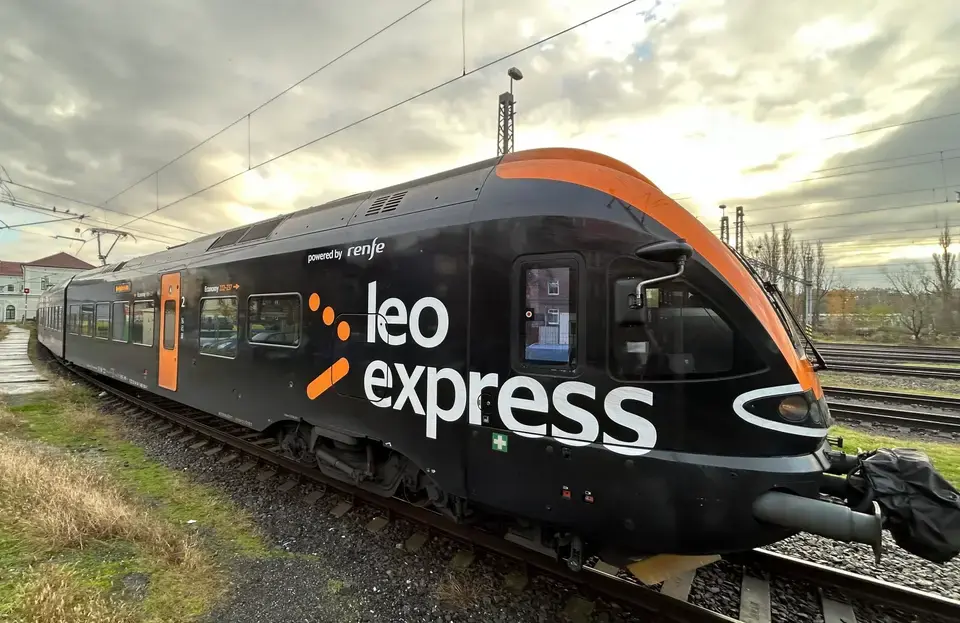 Vlak Leo Express na železničnej trati za súmraku, moderný dizajn, oranžovo-čierna farebná schéma.