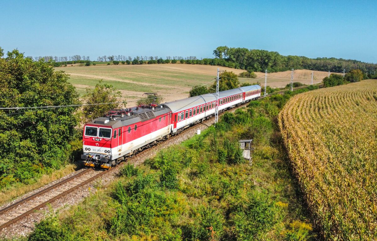 Červený rušeň ťahá osobný vlak cez malebnú slovenskú krajinu medzi poľami a lesmi za jasného dňa.