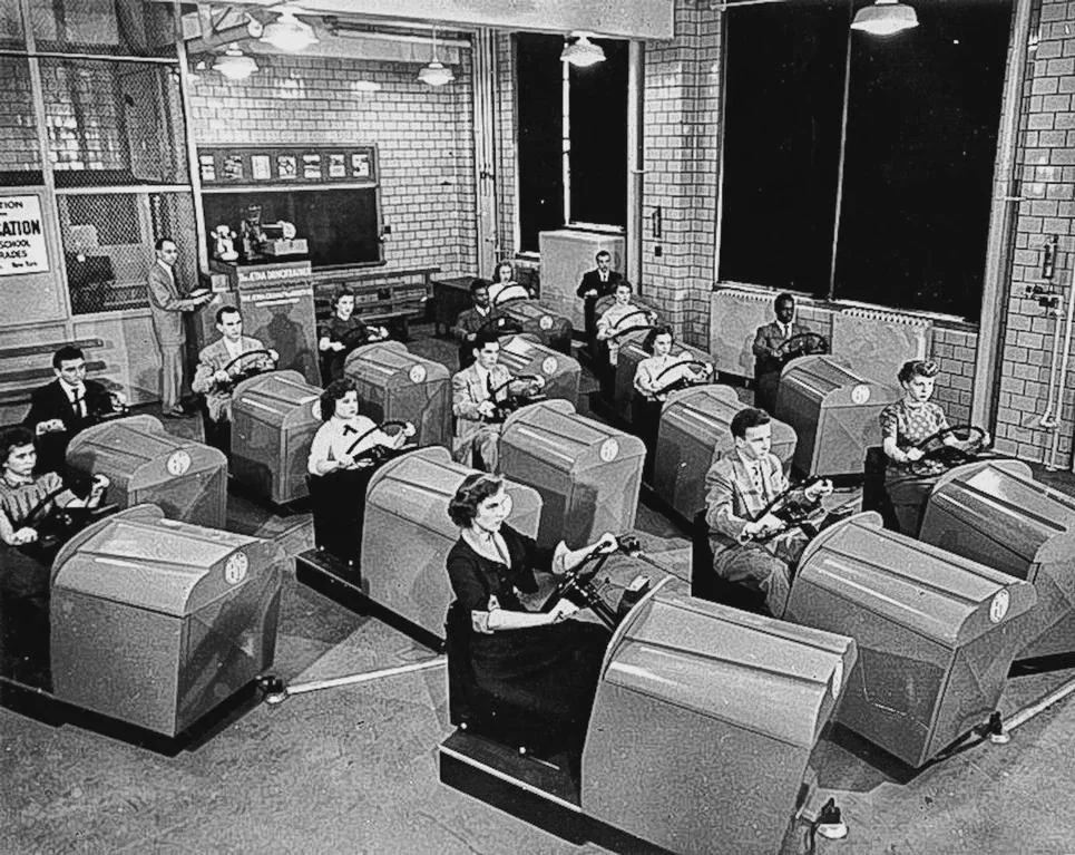 Simulácia autoškola 1950s: Skupina ľudí v triede používa staré cvičné zariadenia na učenie jazdy.
