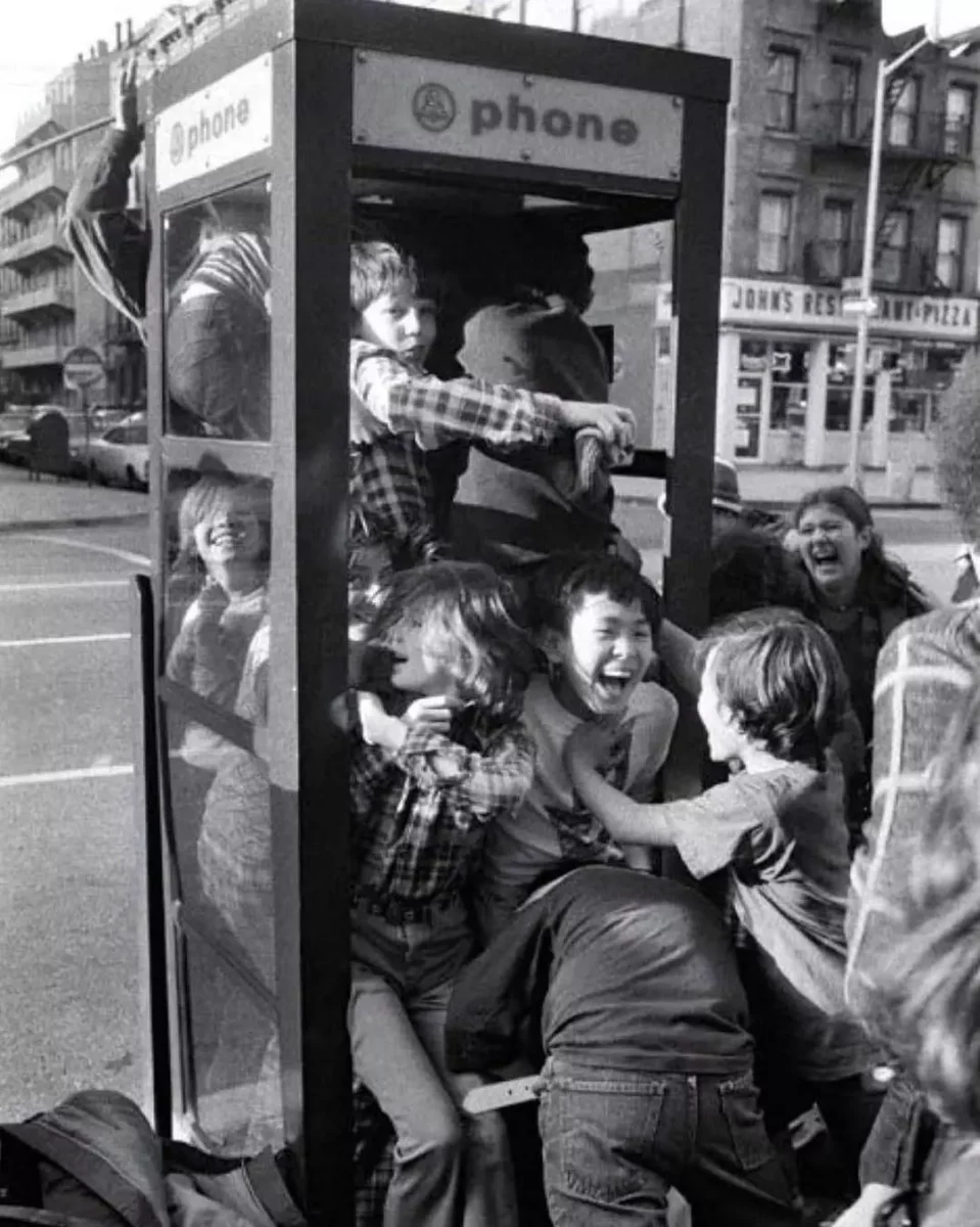 Skupina detí sa smeje a tlačí do telefónnej búdky v meste, stará čiernobiela fotografia.