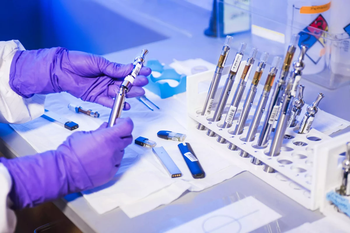 Laboratórny výskum elektronických cigariet, ruky v rukaviciach manipulujú s náplňami a zariadeniami na stole.