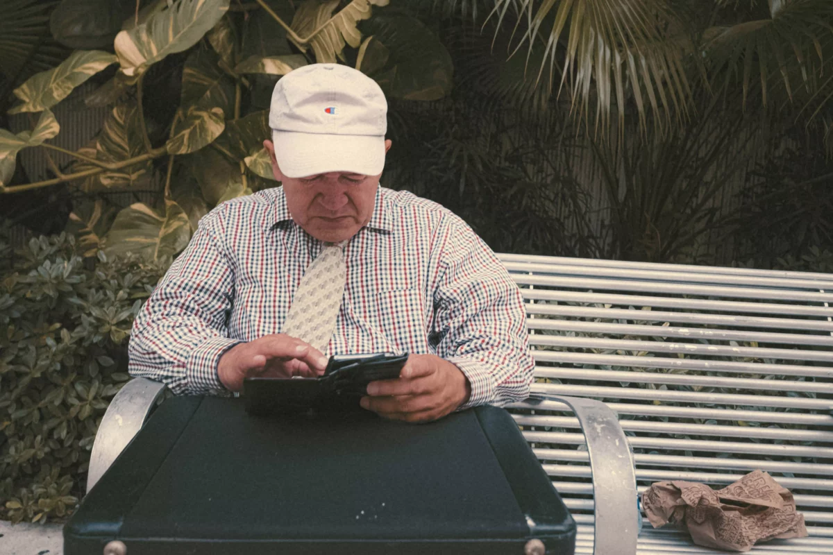Muž sedí na lavičke v klobúku a košeli, používa tablet na čítanie.