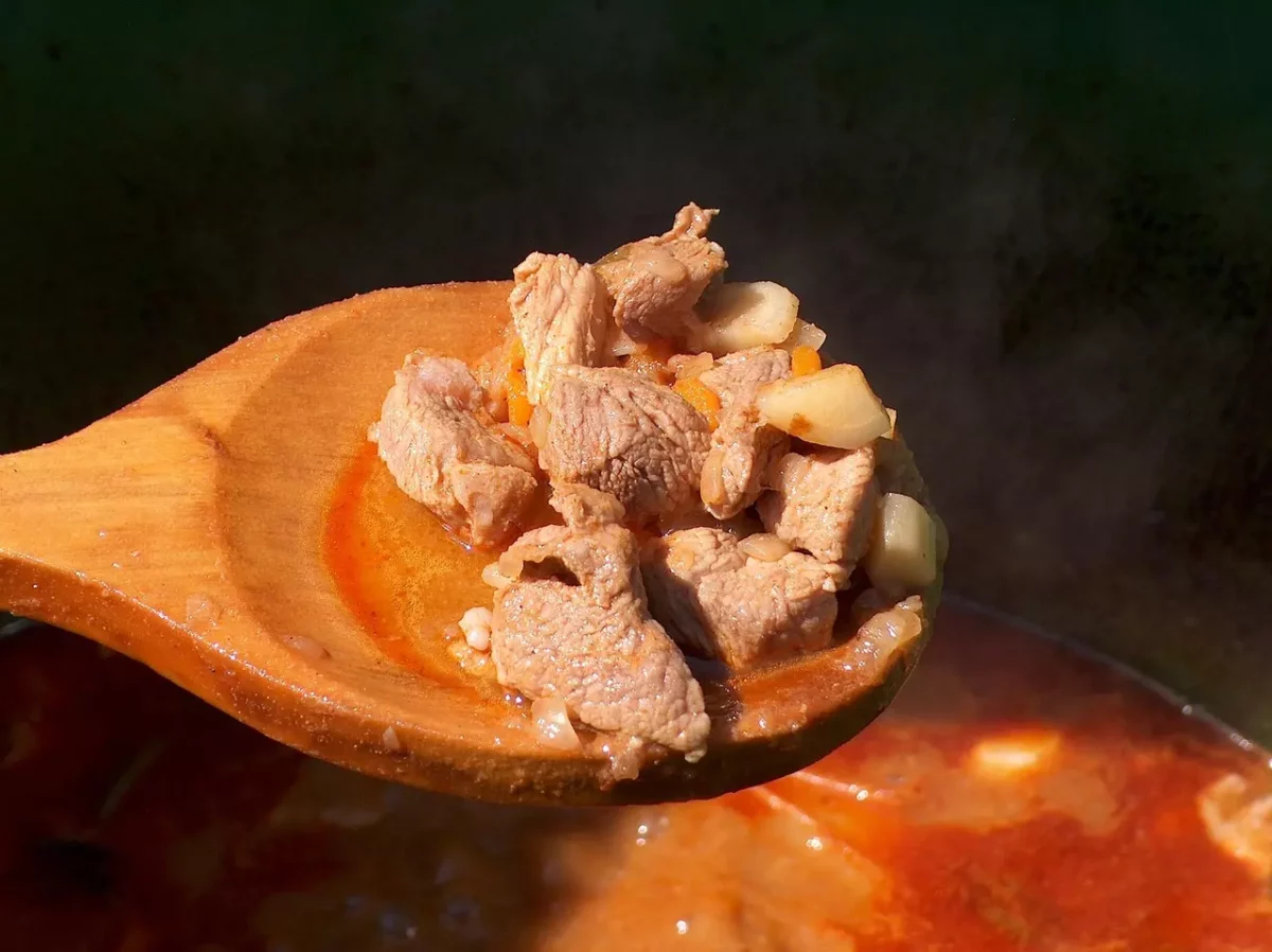 Drevená lyžica s gulášom plným mäsa a zeleniny, nad vriacim kotlom. Chutná delikatesa pripravená na ohni.