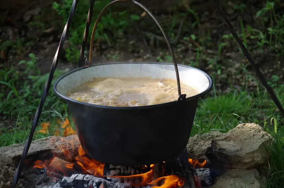 Guláš sa varí v kotlíku nad otvoreným ohňom, vidno drevo a zelené pozadie. Varenie v prírode.