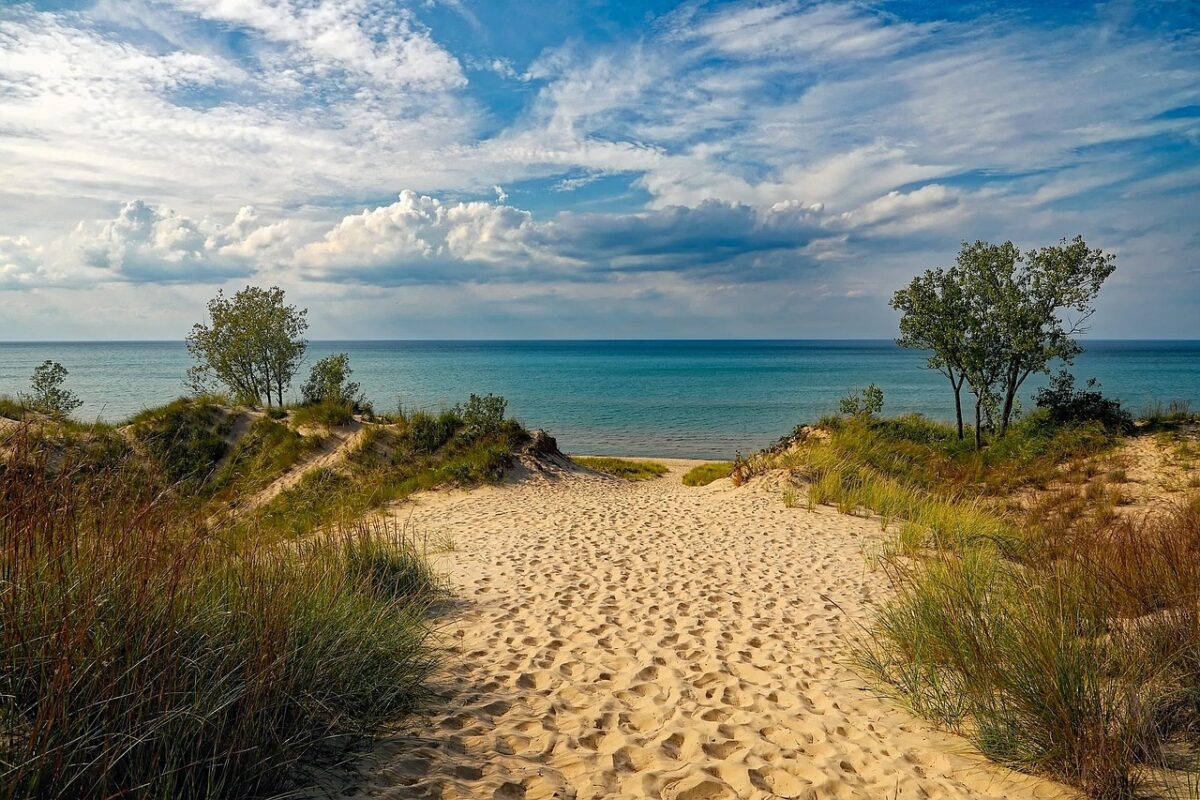Piesočnatá pláž s modrým morom a zelenými dunami pod oblačnou oblohou. Relaxačná scenéria prírody.