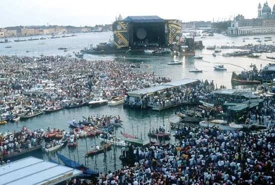 Obrovský dav ľudí a lodí na hudobnom festivale pri vode s pódiom v diaľke.
