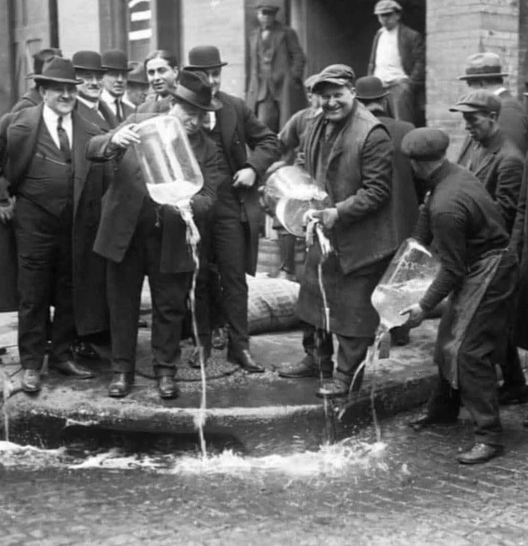 Skupina mužov v klobúkoch vylieva tekutinu z veľkých nádob na ulicu, historická fotografia.
