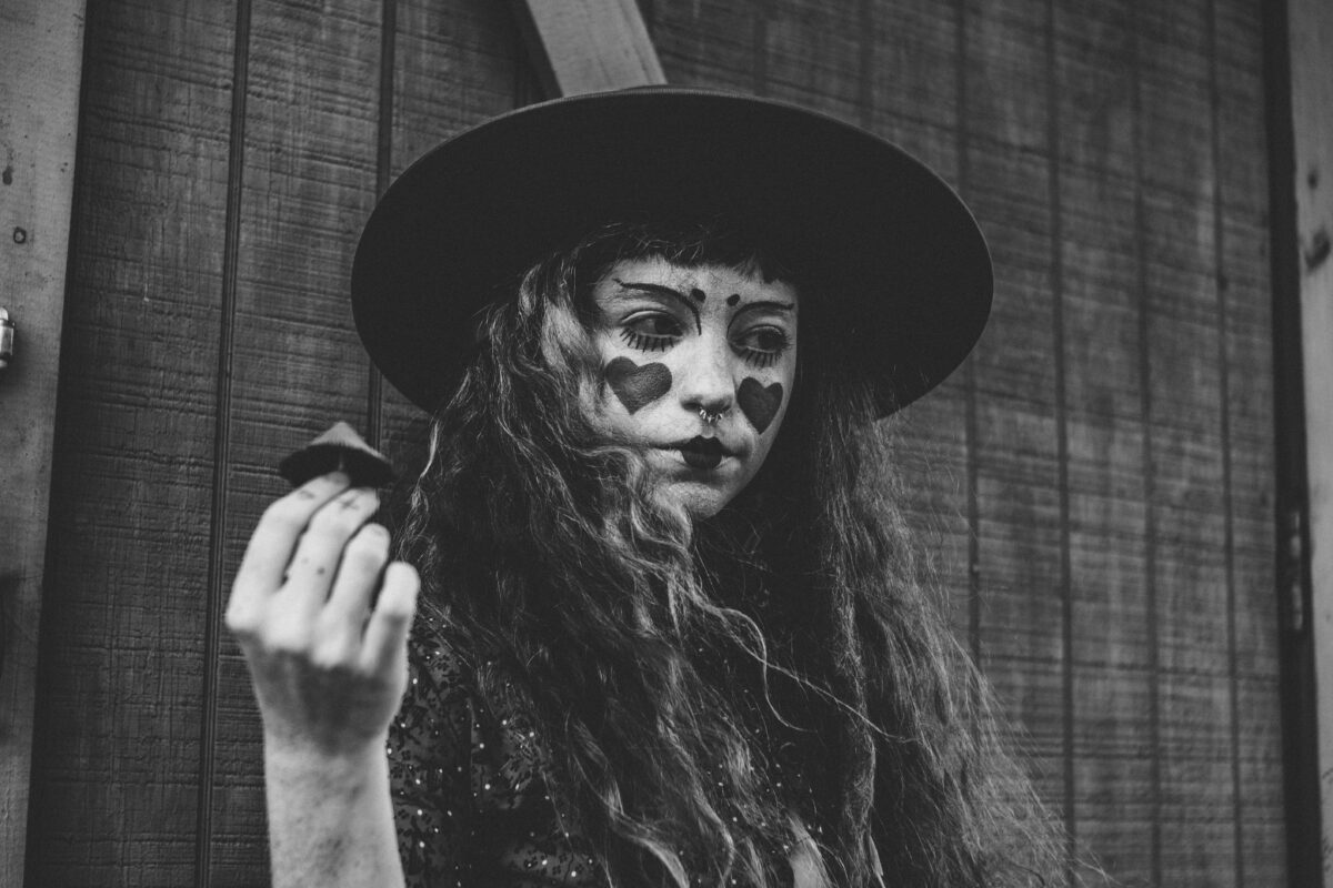 Žena v klobúku s maľovanými srdcami na tvári, drží predmet, čierno-biela fotografia.