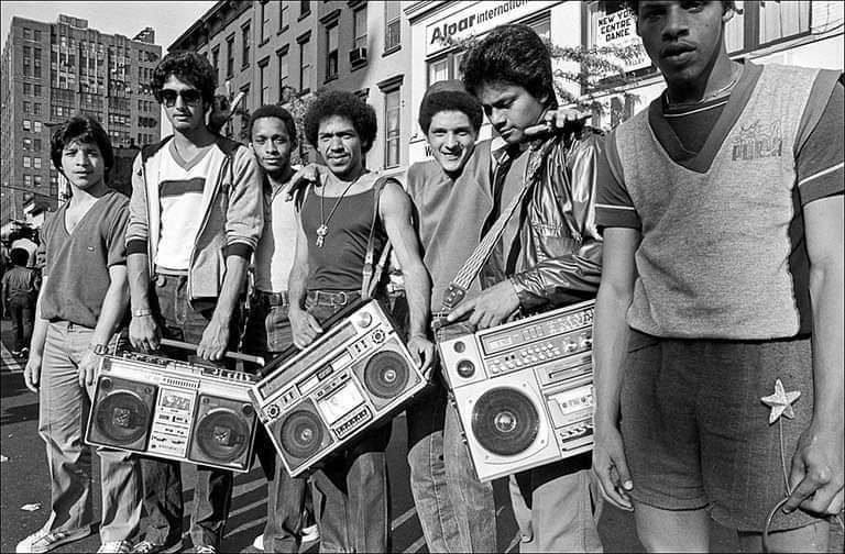 Skupina mladých mužov s boomboxmi na ulici v mestskom prostredí, zachytená v čiernobielej fotografii.