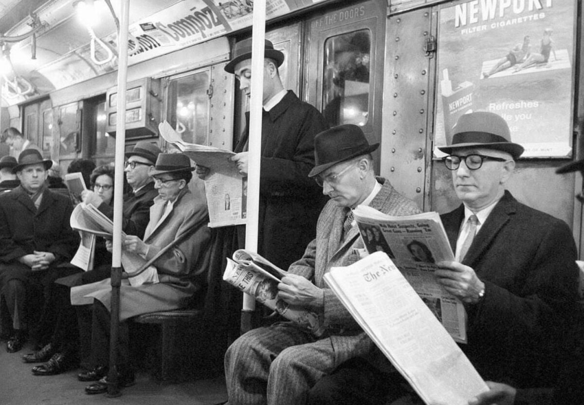 Ľudia v klobúkoch čítajúci noviny v starom vagóne metra. Čiernobiela fotografia.