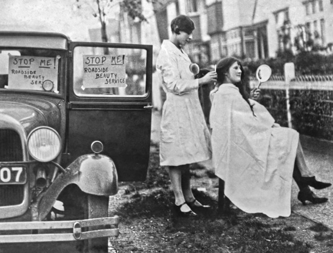 Mobilná kadernícka služba pri ceste v 30. rokoch, žena na stoličke, druhá ju češe, auto s nápisom Roadside Beauty Service.