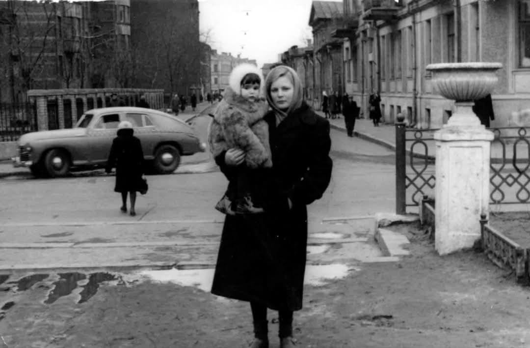 Matka s dieťaťom v historickej ulici, zimné oblečenie, autá a ľudia v pozadí.