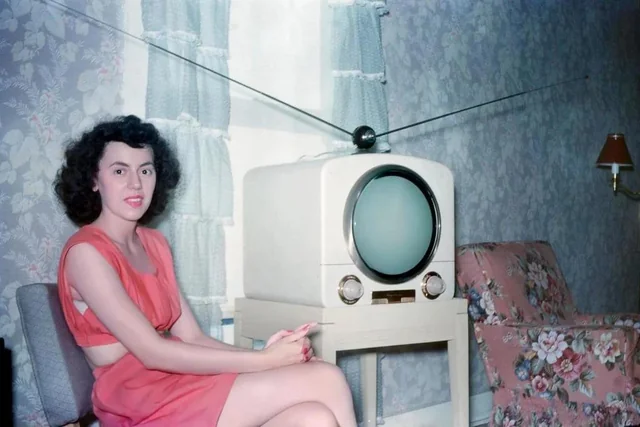 Žena sedí vedľa retro televízora, oblečená v červenom odeve, v miestnosti s kvetinovým nábytkom a závesmi.