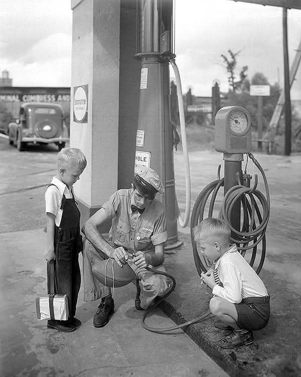 Muž opravuje pumpu na čerpacej stanici, zatiaľ čo dvaja chlapci ho pozorujú so záujmom. Retro atmosféra.