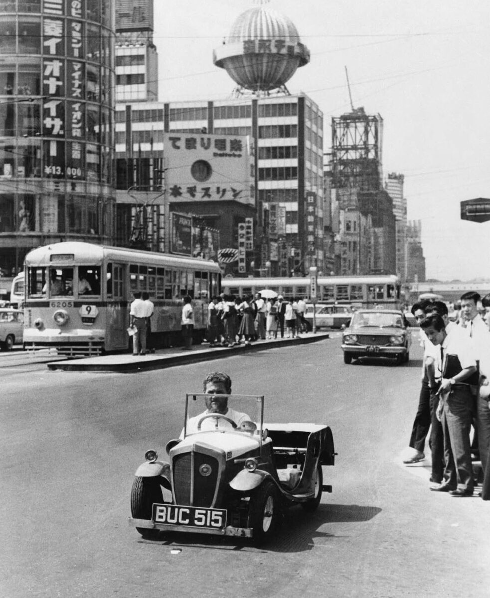 Muž v malom aute na rušnej japonskej ulici s obchodmi a električkou v pozadí. Retro atmosféra ázijského veľkomesta.