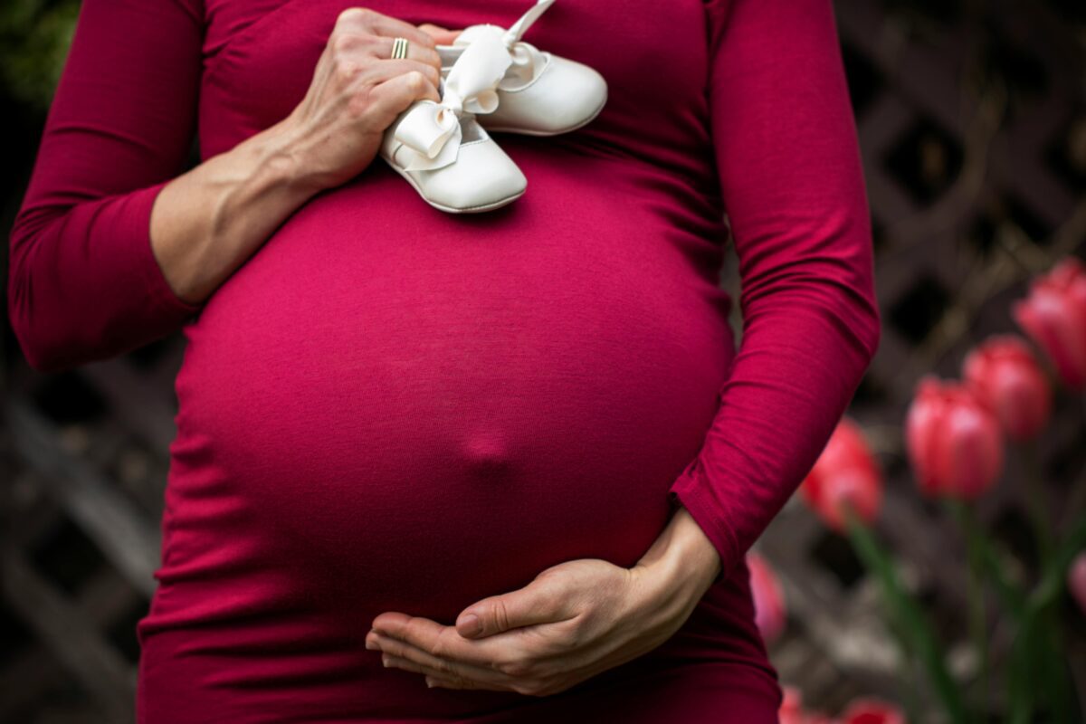 Tehotná žena v červených šatách držiaca detské topánky na brušku, v pozadí rozmazané červené kvety.