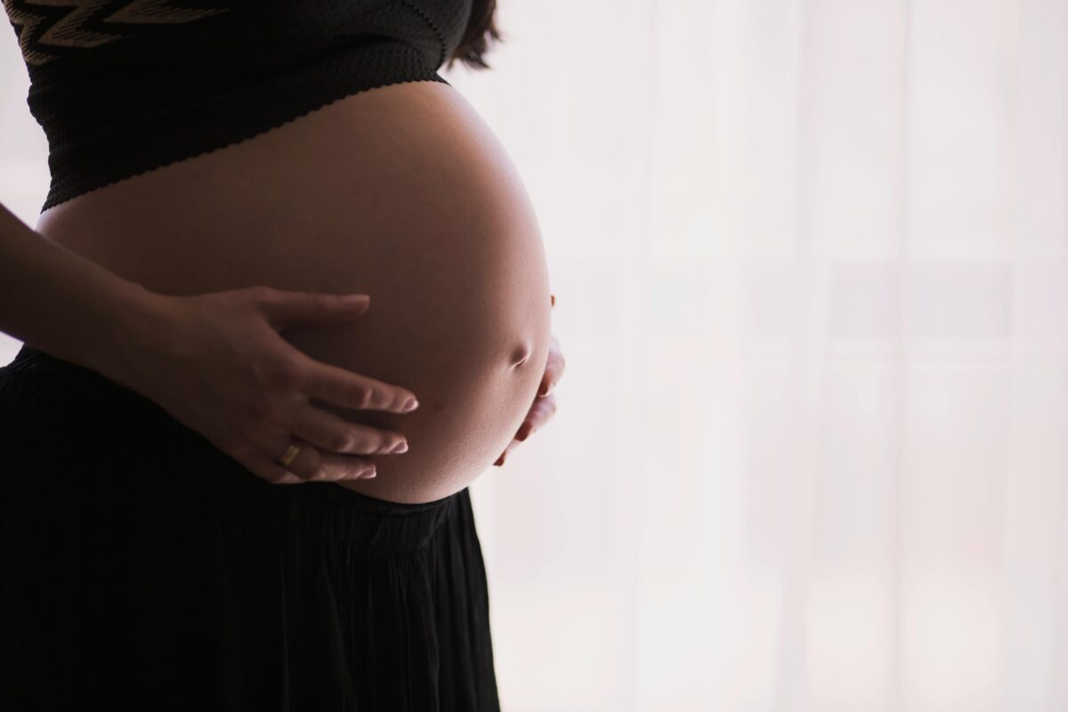 Tehotná žena držiaca sa za brucho, stojaca pri okne s jemným osvetlením v pozadí.