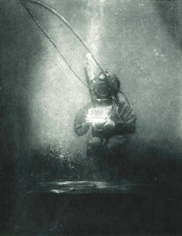 Historický potápač s helmou drží svetlo, pod vodou, obklopený hmlistým svetlom.