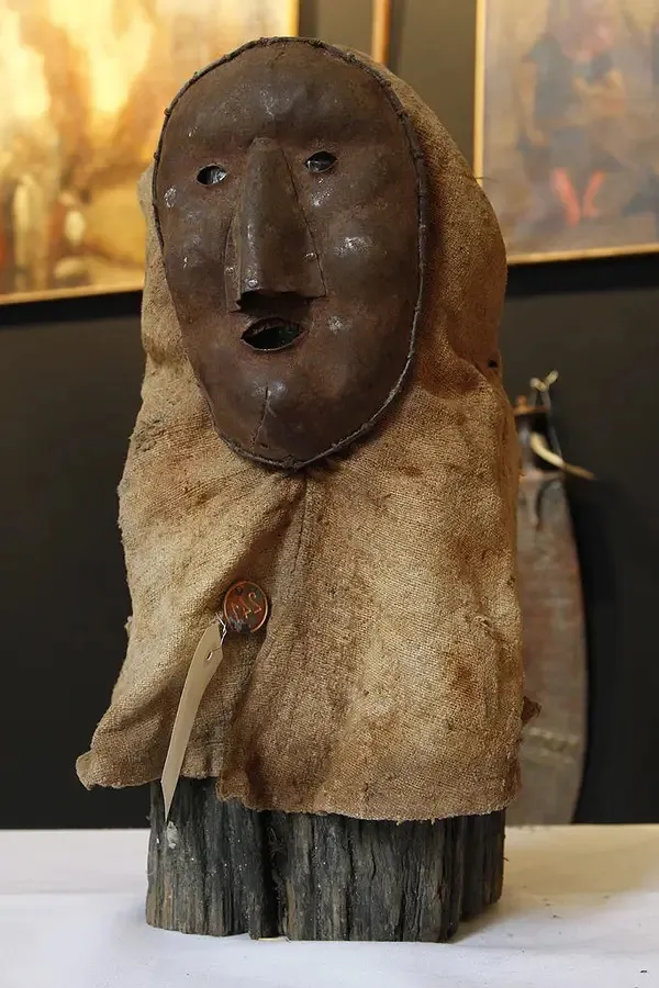 Drevená socha s maskou z látky, vystavená v múzeu na podstavci, obklopená historickými obrazmi.