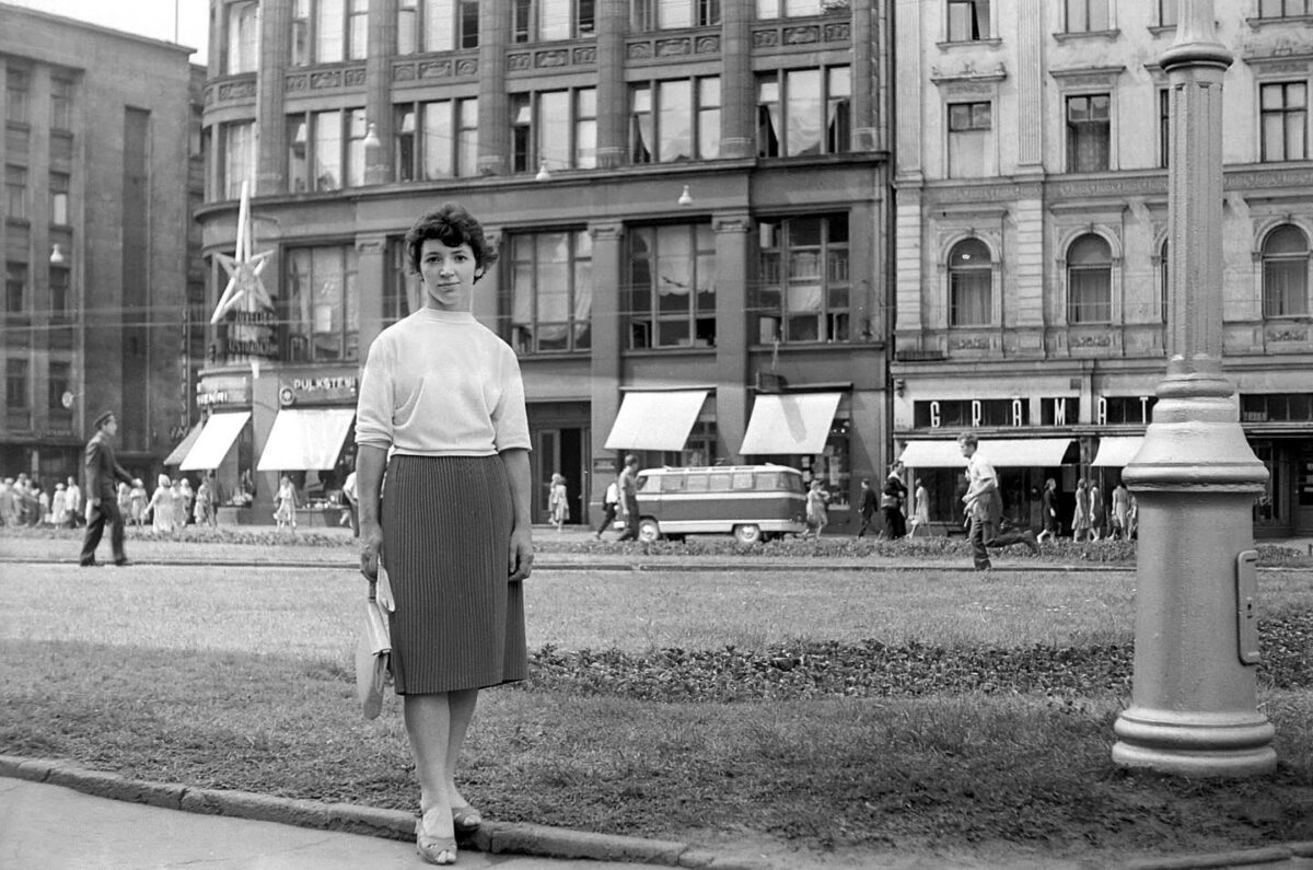 Žena stojí na ulici pred historickou architektúrou, okolo idú ľudia. Vintage čiernobiela fotografia mesta.