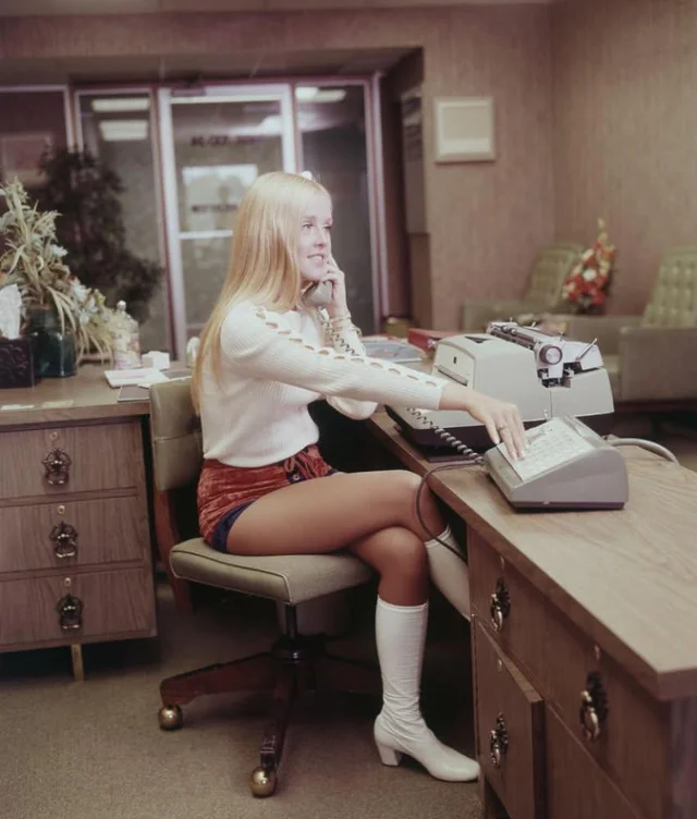 Žena v kancelárii 70. rokov telefonuje a pracuje s písacím strojom, sedí za retro dreveným stolom.