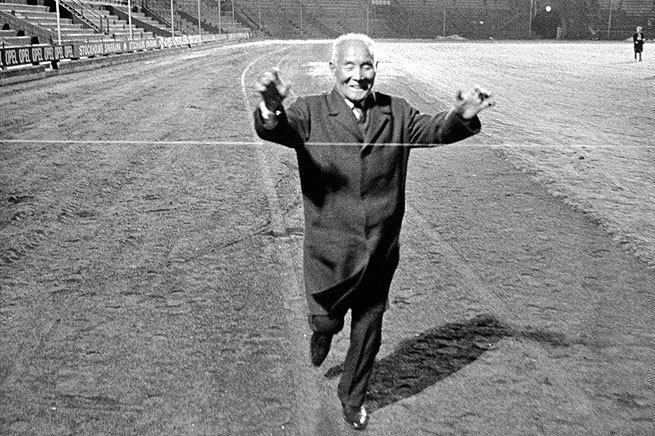 Starší muž v obleku prebieha cieľovou páskou na atletickej dráhe, večerná atmosféra.