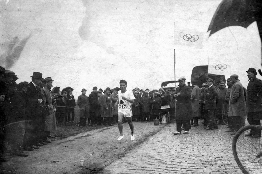 Bežec v pretekoch obklopený divákmi, historická atmosféra s olympijskými vlajkami na pozadí.