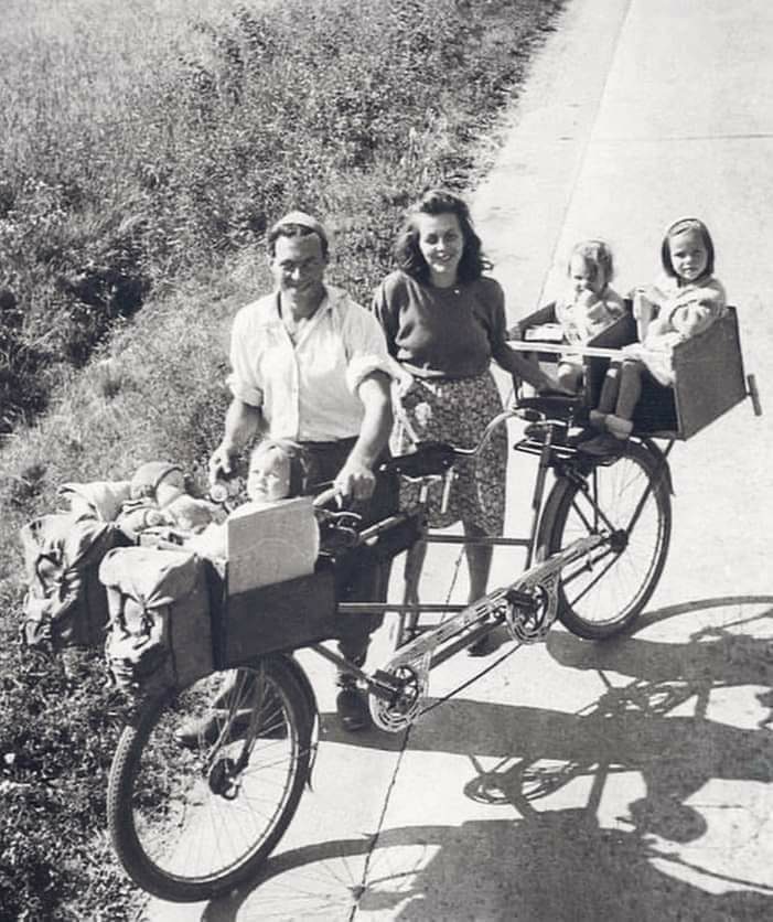 Rodina počas cyklistického výletu na tandeme so štyrmi deťmi v drevených nosičoch, čiernobiela historická fotografia.