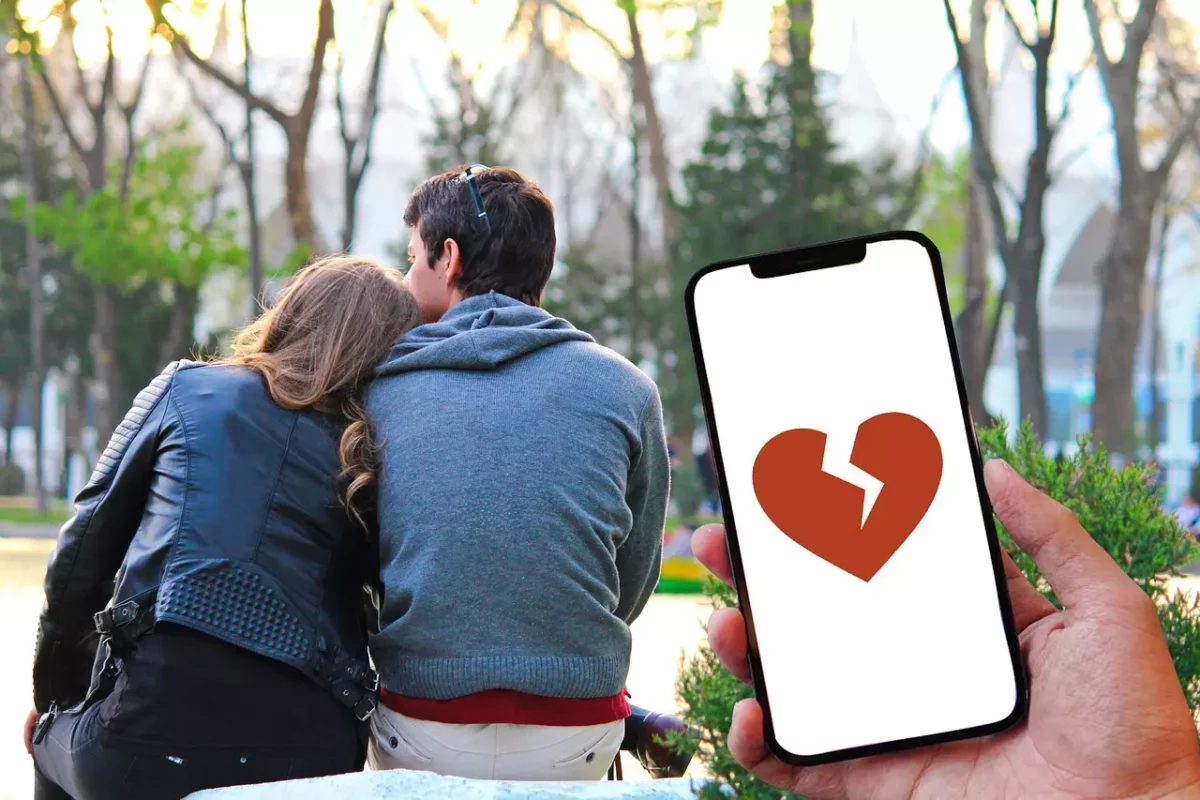 Pár sediaci v parku, smartphone s obrázkom zlomeného srdca, symbol narušeného vzťahu.