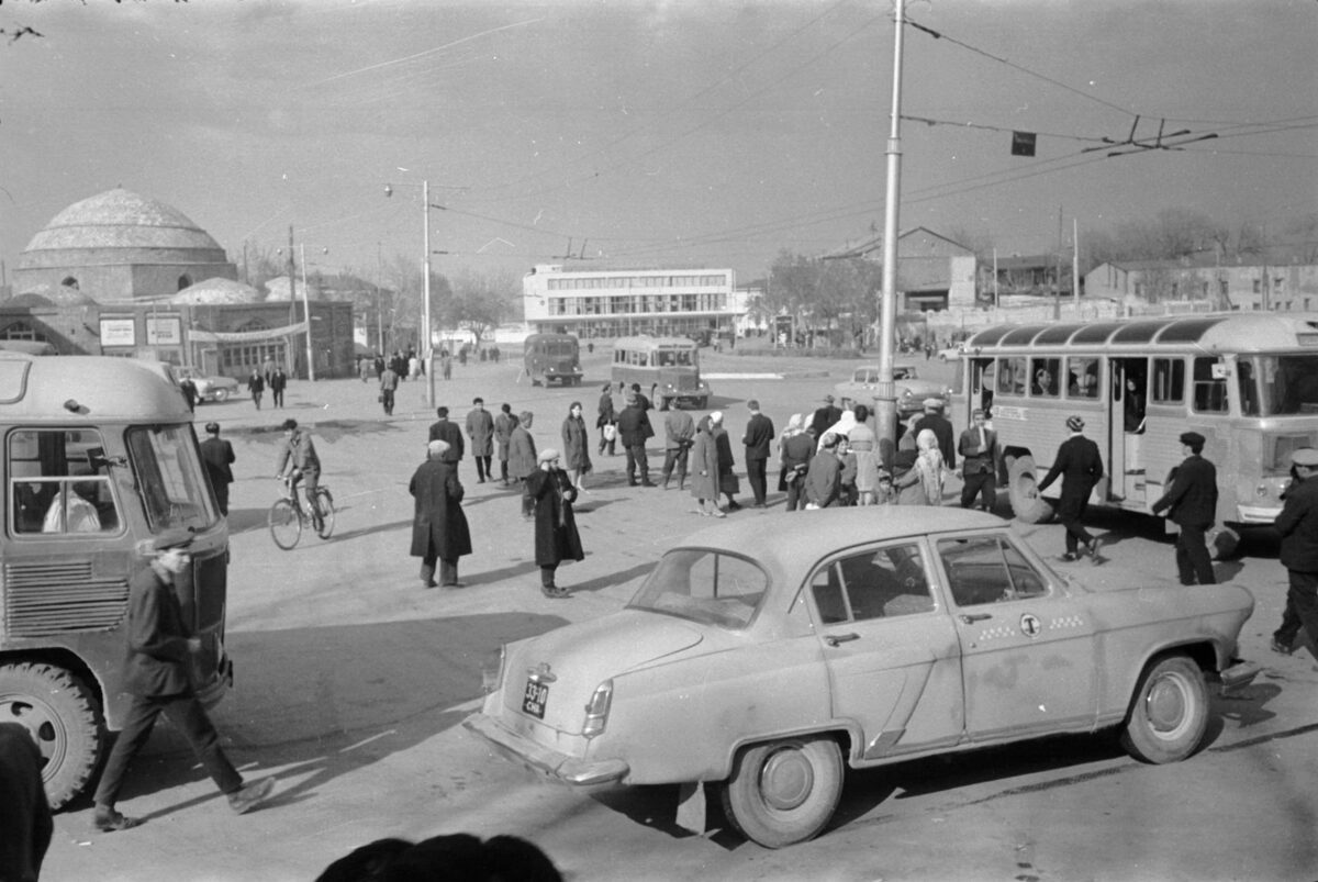 Čiernobiela fotografia rušného mestského námestia s autami, chodcami a historickými budovami v pozadí.