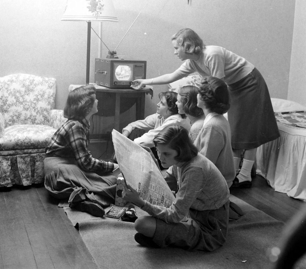 Skupina dievčat sleduje televíziu, jedna číta noviny v retro obývačke, 50. roky.
