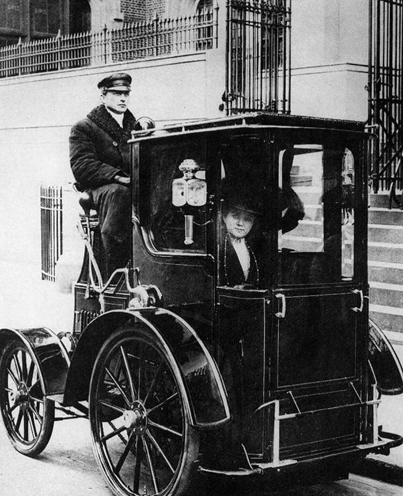 História automobilizmu: Staromódny automobil s vodičom a elegantnou dámou na mestskej ulici.