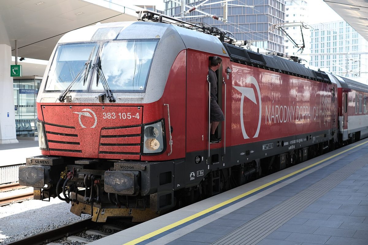 Červená lokomotíva Siemens Vectron na stanici, vedľa stojí osoba. Moderná železničná doprava v meste.