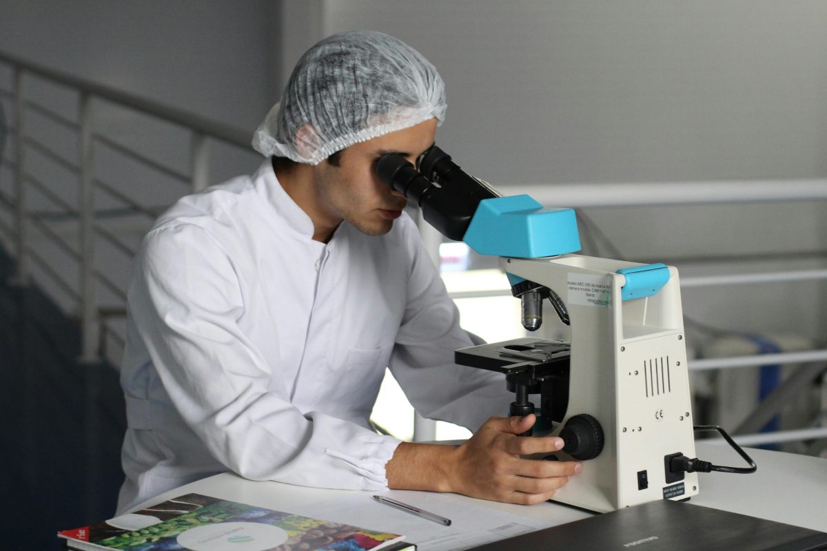 Výskumník v laboratóriu skúmajúci vzorky pod mikroskopom, oblečený v bielej laboratórnej blúze.