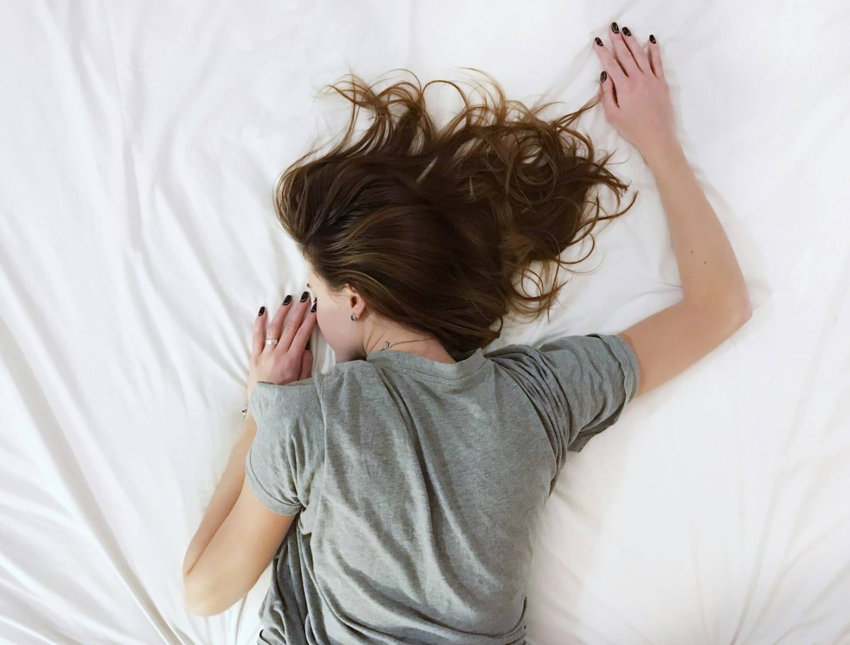 Žena relaxuje na bielej posteľnej bielizni, oblečená v šedom tričku, s rozviatymi vlasmi.
