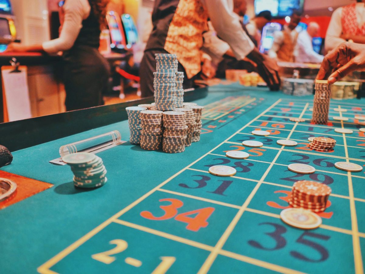 Ruleta v kasíne s farebnými žetónmi na stole, hráči v pozadí dávajú stávky.