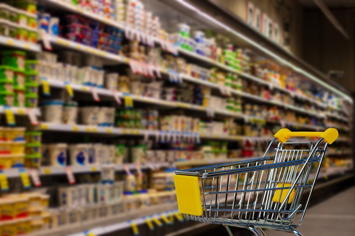 Nákupný košík v supermarkete, poličky plné potravín v pozadí.