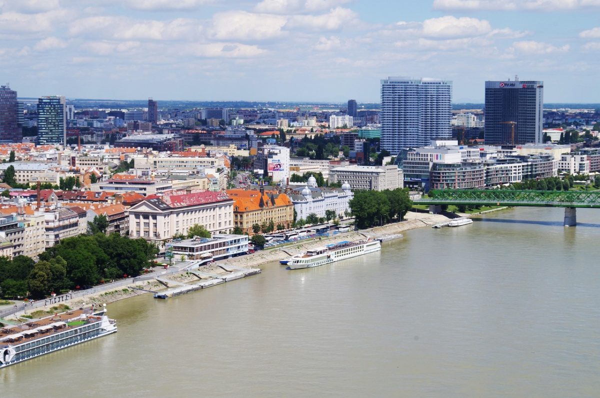 Pohľad na Bratislavu pri rieke s modernými budovami a plavidlami na Dunaji za slnečného dňa.